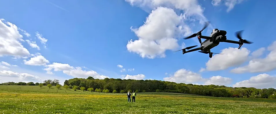 Drone DJI en vol au-dessus d’une prairie fleurie, deux télépilotes debout au loin, ciel bleu nuageux
