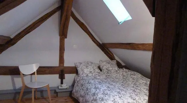 Chambre de l’appartement duplex sous les poutres apparentes, lit double, chaise blanche et fenêtre de toit
