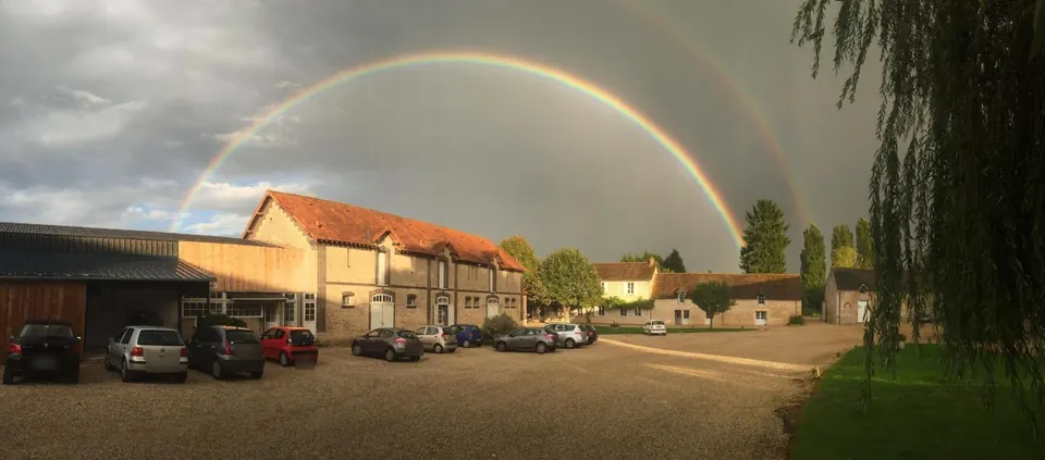 Cour du campus de Marcq : bâtiment de ferme en pierre et brique au toit de tuiles, hangar et voitures garées, sous un arc-en-ciel